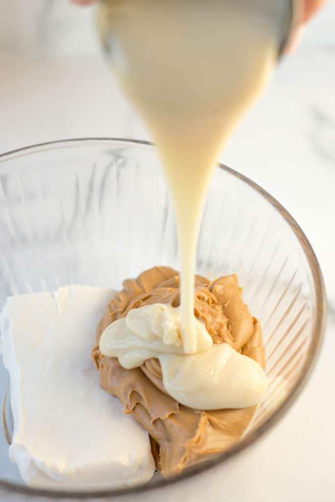 Condensed milk being poured into a clear bowl with a brick of cream cheese and some peanut butter