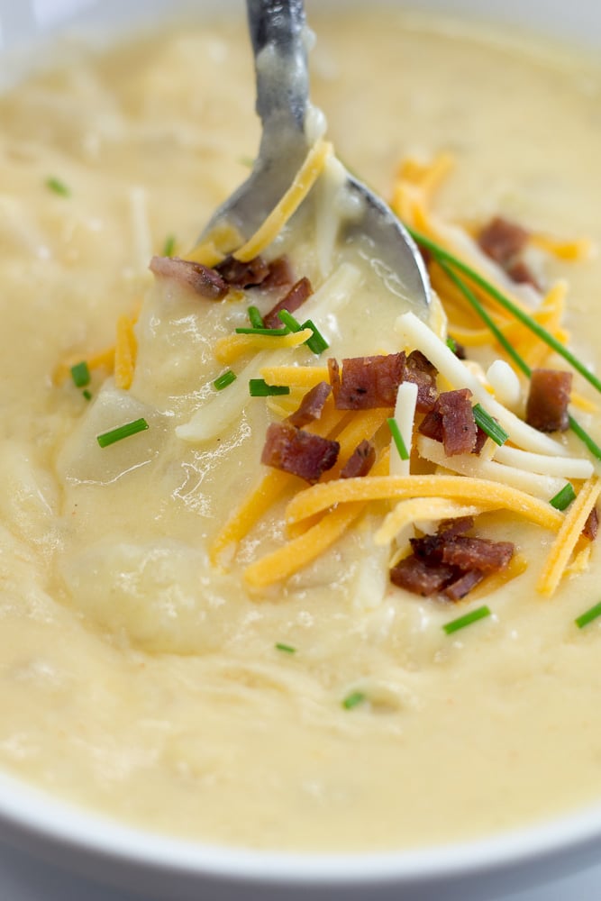 A spoon in a bowl of potato soup garnished with chopped bacon, shredded cheddar cheese, and chives