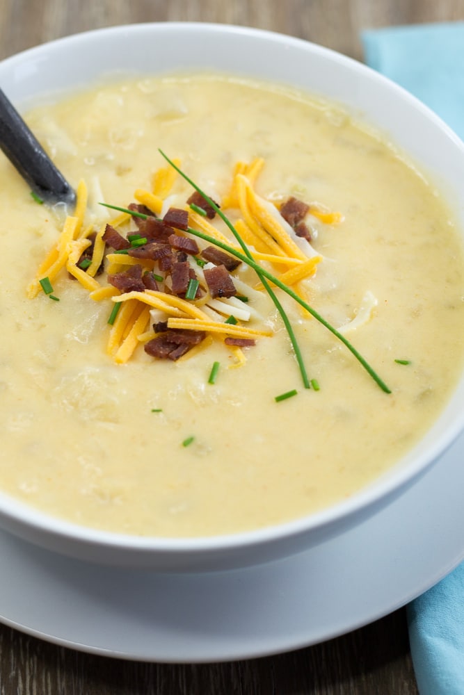 A white bowl of potato soup garnished with bacon, shredded cheddar cheese, and chives