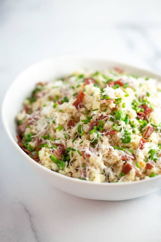 A white bowl filled with cauliflower rice, bacon, peas, parmesan cheese, and chives