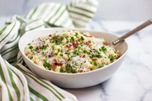 A white bowl filled with cauliflower rice, bacon, peas, parmesan cheese, and chives next to a green striped napkin
