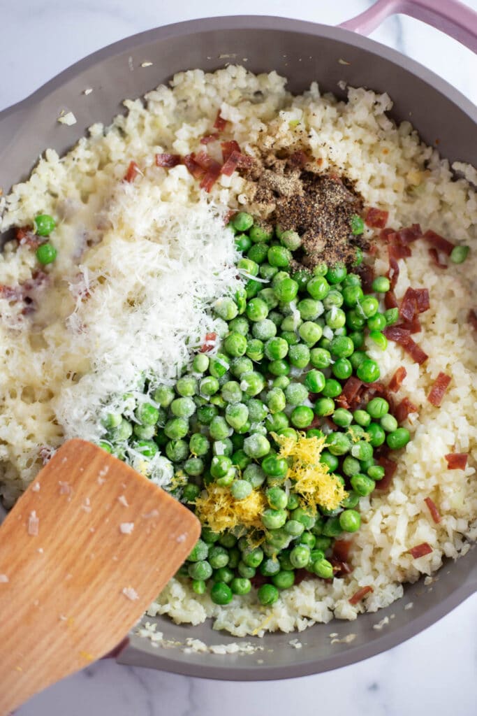 Overhead view of a skillet with cauliflower rice, bacon, peas, parmesan cheese, and lemon zest