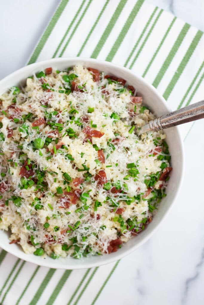 Overhead view of a white bowl filled with cauliflower rice, bacon, peas, parmesan cheese, and chives next to a green striped napkin
