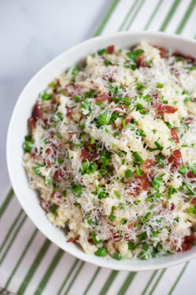 Overhead view of a white bowl filled with cauliflower rice, bacon, peas, parmesan cheese, and chives next to a green striped napkin