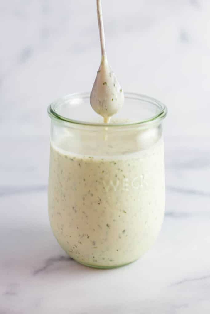 A spoon coated with ranch dressing over the top of a clear jar of ranch dressing