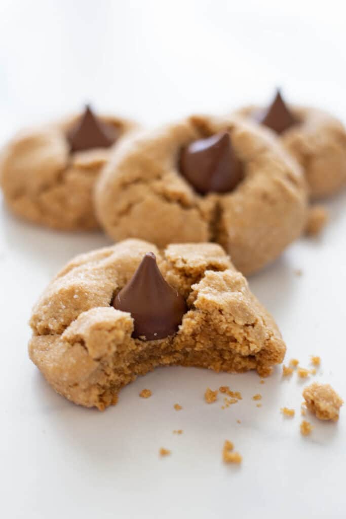 A peanut butter kiss cookie missing a bite next to cookie crumbs with more cookies in the background