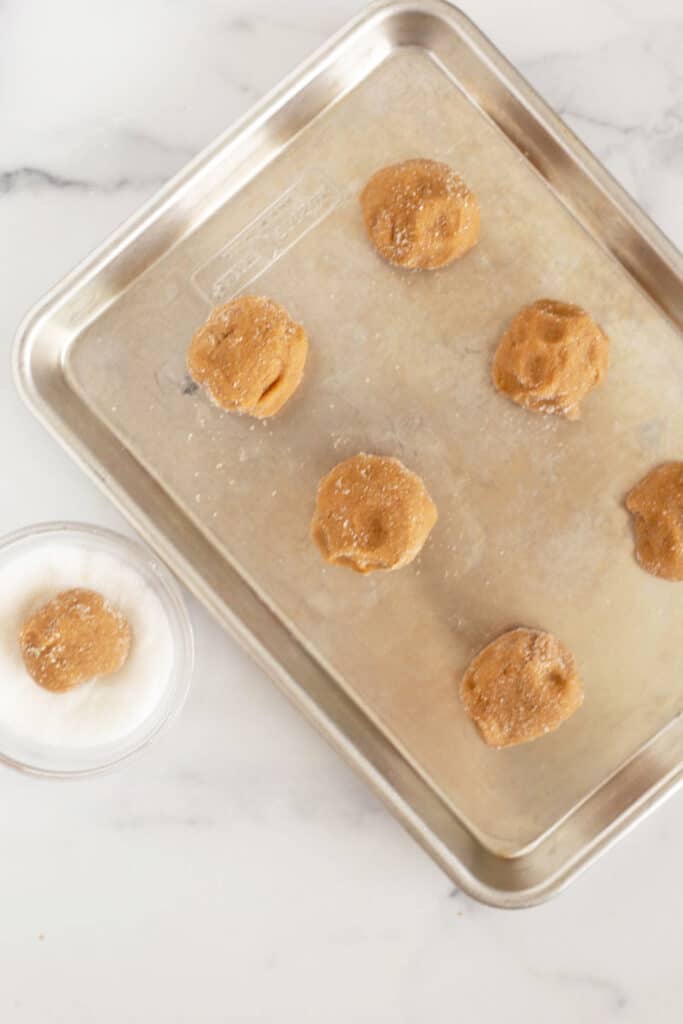 Overhead view of uncooked, balled cookie dough on a baking sheet next to a small bowl of white sugar