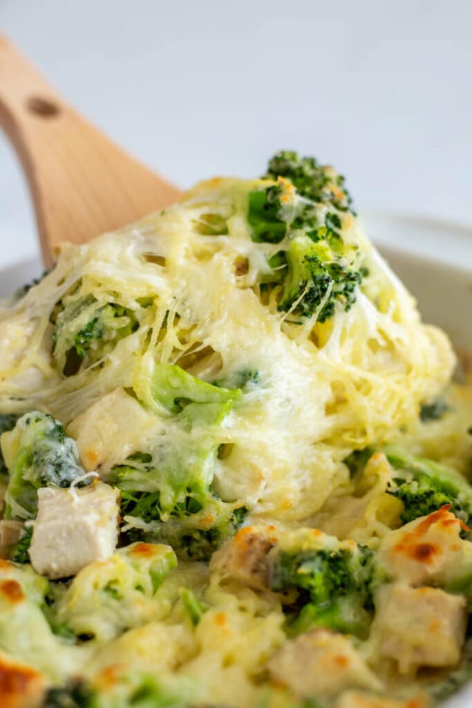 Wooden spoon scooping cheesy chicken, broccoli spaghetti squash casserole from a white baking dish