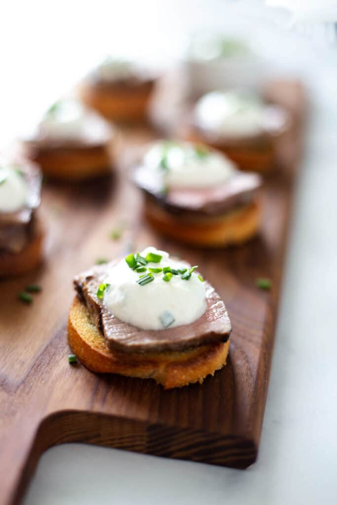 Crostini topped with steak and white sauce on a wood cutting board