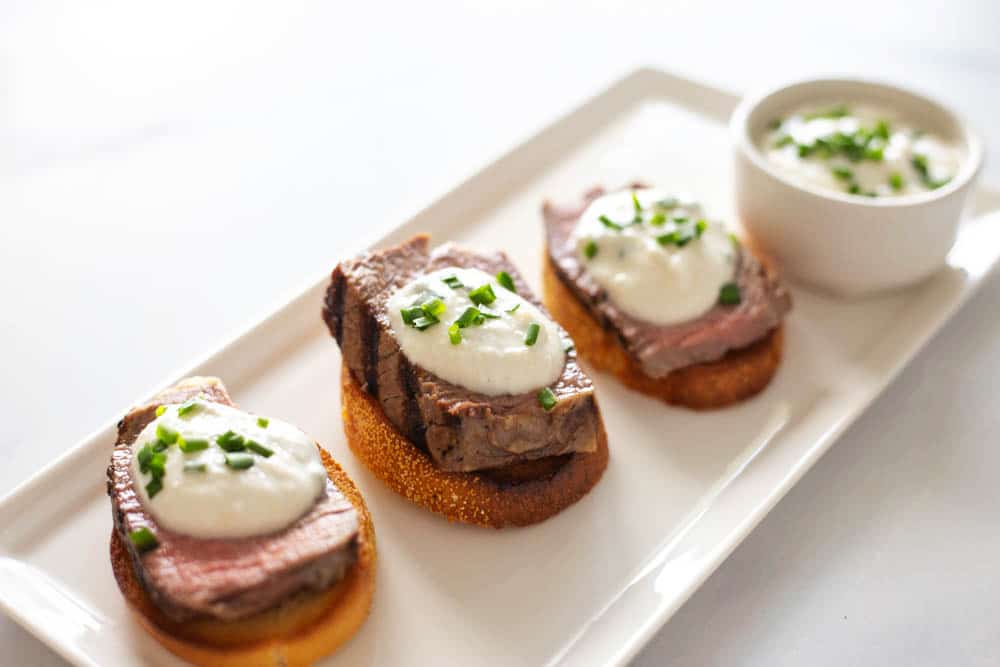 Crostini topped with steak and a white sauce on a rectangular plate