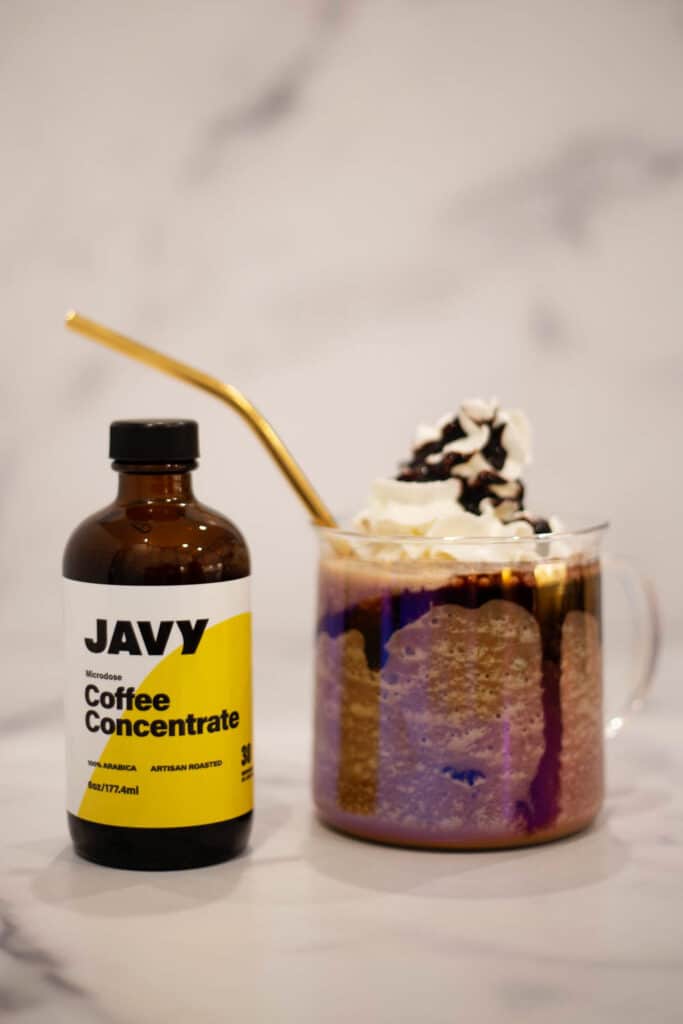 Clear coffee mug filled with chocolate frappe topped with whipped cream and a gold straw next to a bottle of javy coffee concentrate