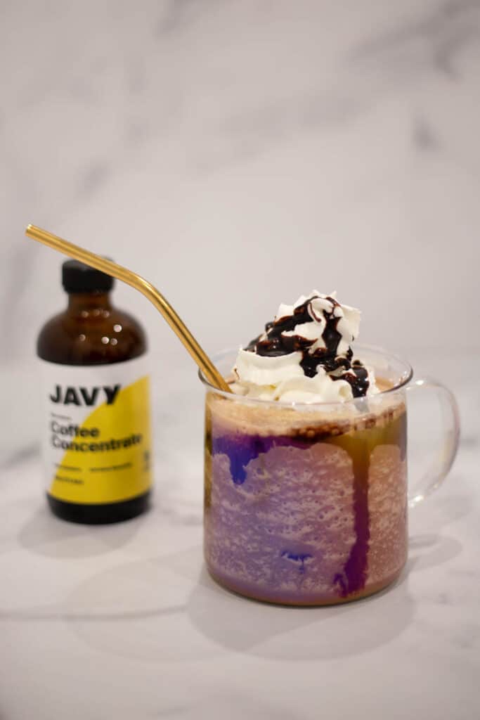 Clear coffee mug filled with chocolate frappe topped with whipped cream and a gold straw next to a bottle of javy coffee concentrate