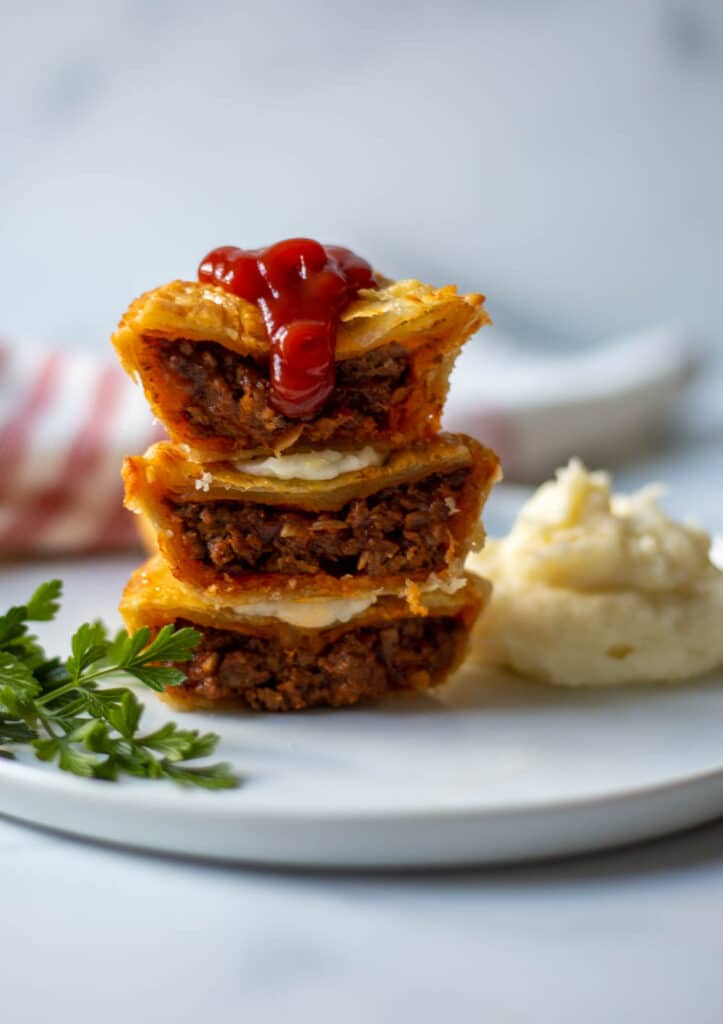 Stack of 3 Australian meat pies on a white plate next to a pile of mashed potatoes garnished with sprigs of parsley