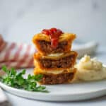 Stack of 3 meat pies on a white plate next to a pile of mashed potatoes garnished with sprigs of parsley